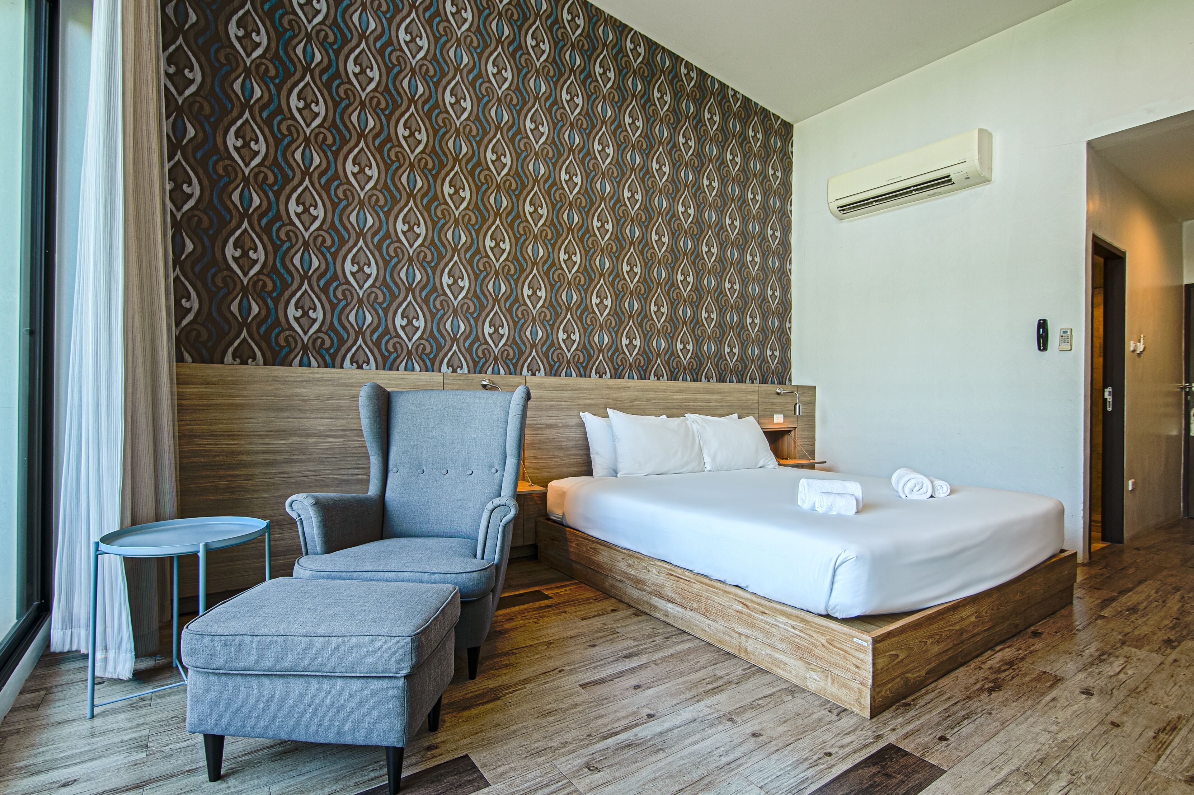 Una habitación de hotel con una cama, un sillón gris, una mesita y un papel tapiz estampado.
