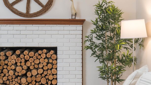 Ein gemütliches Wohnzimmer mit Kamin aus weißem Mauerwerk, einer großen Friedenssymbol-Dekoration, einem Holzstapel, einem weißen Sofa mit kariertem Überwurf, einer hohen Zimmerpflanze und einer Stehlampe.