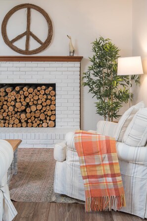 A cozy living room with a white brick fireplace, a large peace sign decoration, a stack of logs, a white sofa with a plaid throw blanket, a tall green plant, and a floor lamp.