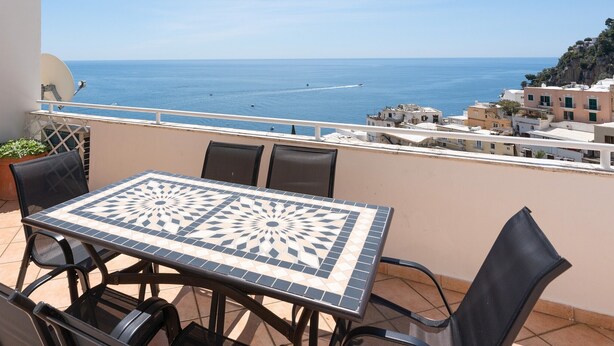 Un balcón con mesa y sillas de cerámica, con vistas a la costa, edificios y el mar.