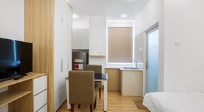 A compact hotel room with a TV, a small dining table, a chair, a kitchenette, and a bed.