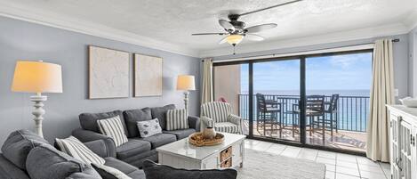 A living room with grey sofas, a white coffee table, and a ceiling fan. There is a large sliding glass door leading to an outdoor area with a view of the ocean.