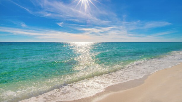Une plage à l’eau turquoise et limpide, un rivage sablonneux et un soleil éclatant dans le ciel.