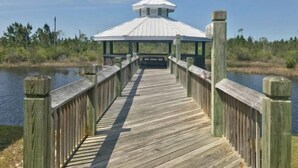 A wooden boardwalk leading to a white gazebo with a peaked roof, surrounded by water and greenery.