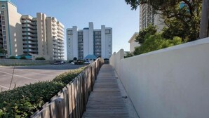 A wooden boardwalk leading to a parking area with multiple cars, surrounded by a low wall and greenery.