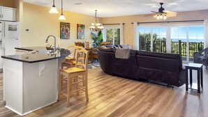 A modern kitchen with a central island, wooden dining chairs, a dark sofa, and a balcony with a view. - Fall is spectacular on the Cape! Book your trip now (Port St Joe)