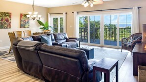 A spacious living room with a leather sectional sofa, a dining area with wicker furniture, and large sliding glass doors leading to a balcony with a view of the ocean. - Fall is spectacular on the Cape! Book your trip now (Port St Joe)