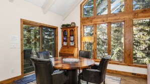 A dining area with a wooden table and chairs, large windows with a forest view, and a wooden cabinet with decorative items. - Glen Lyon Lodge | Spacious 5BR Vail Home, Cascade Lift #20 (Vail)