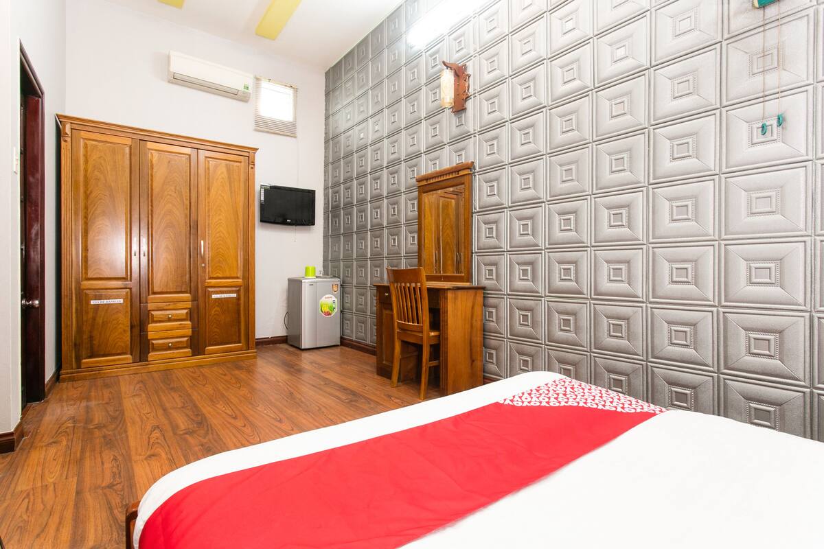 Habitación de hotel con armario de madera, escritorio y silla, cama con colcha roja y blanca, y pared con diseño geométrico.