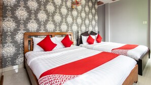 Quarto de hotel com duas camas, cada uma com roupa de cama branca e detalhes vermelhos, papel de parede estampado e um ventilador de teto.