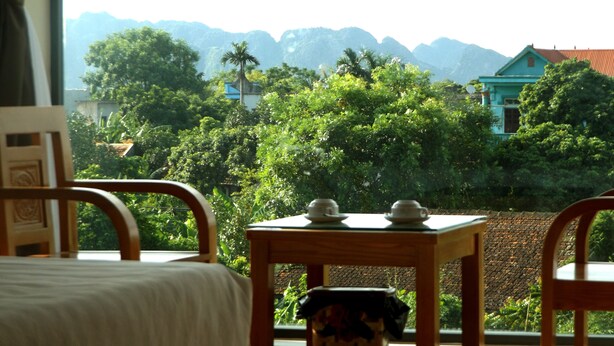 A room with a bed, a table with two cups, and a view of lush greenery and mountains. - Ninh Binh Riverside Homestay 2 (Ninh Binh)