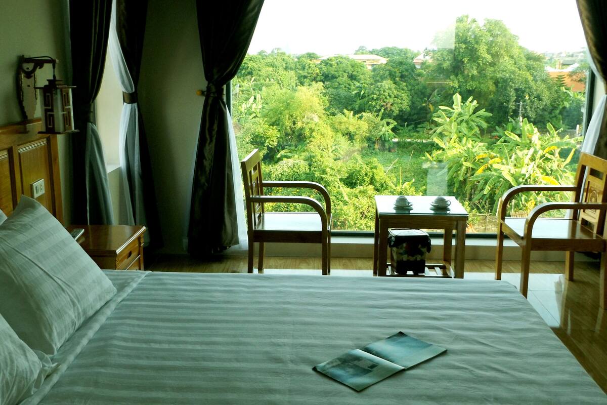 A hotel room with a bed, a small table with chairs, and a view of a lush garden.