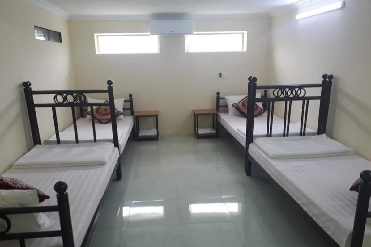 A room with four single beds, each with a wooden frame and white bedding. There are two small bedside tables with lamps, and a window above the beds.