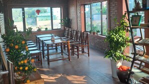 A spacious room with a brick wall, large windows, and a wooden table with chairs. There are potted plants and a ladder against the wall. - Ninh Binh Riverside Homestay 2 (Ninh Binh)