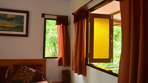 A room with a bed, a window with curtains, a painting on the wall, and a view of greenery outside.