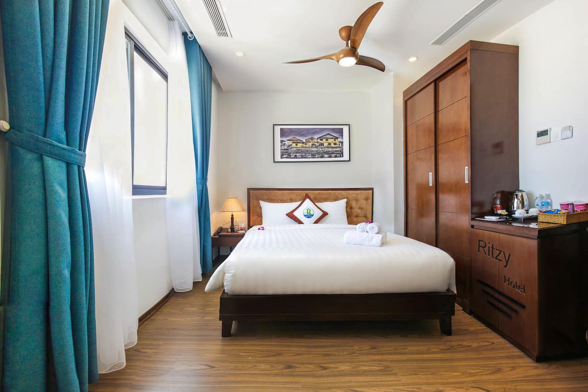 A hotel room with a large bed, a wooden wardrobe, a ceiling fan, a nightstand with a lamp, and a framed picture on the wall.