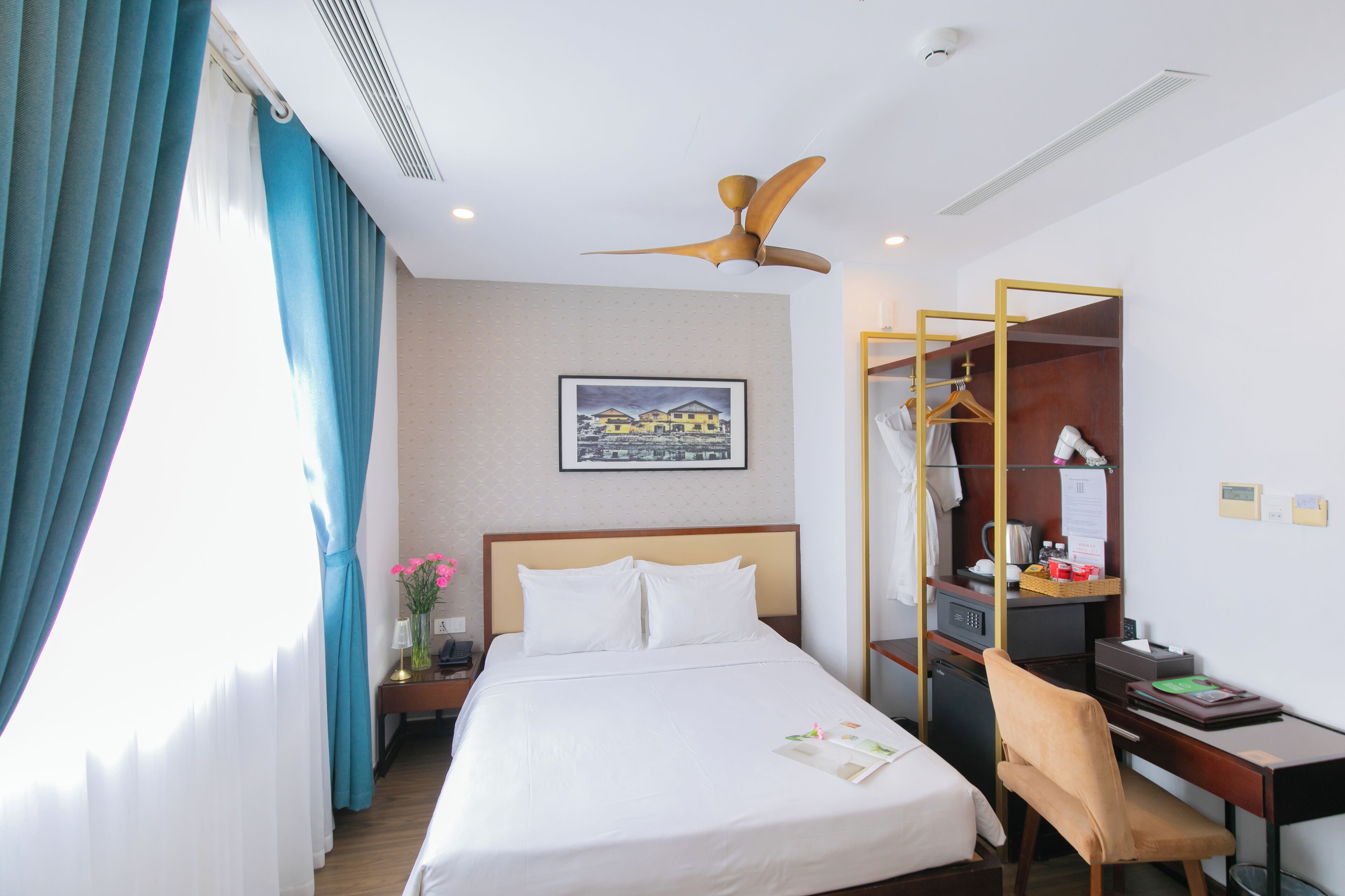 Una camera d'albergo con un letto, una scrivania, una sedia, un armadio, una ventola a soffitto e un quadro appeso al muro.