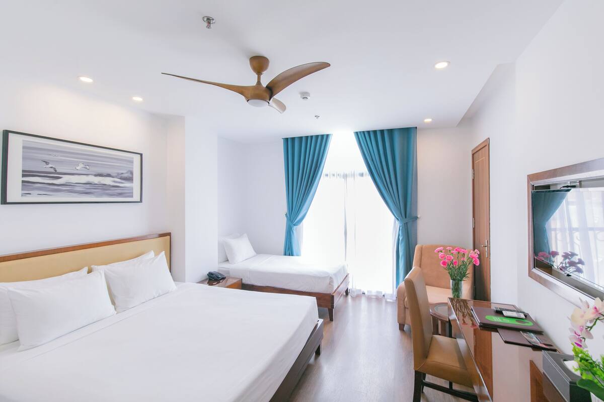 A hotel room with two single beds, a desk with a chair, a ceiling fan, and a window with curtains.