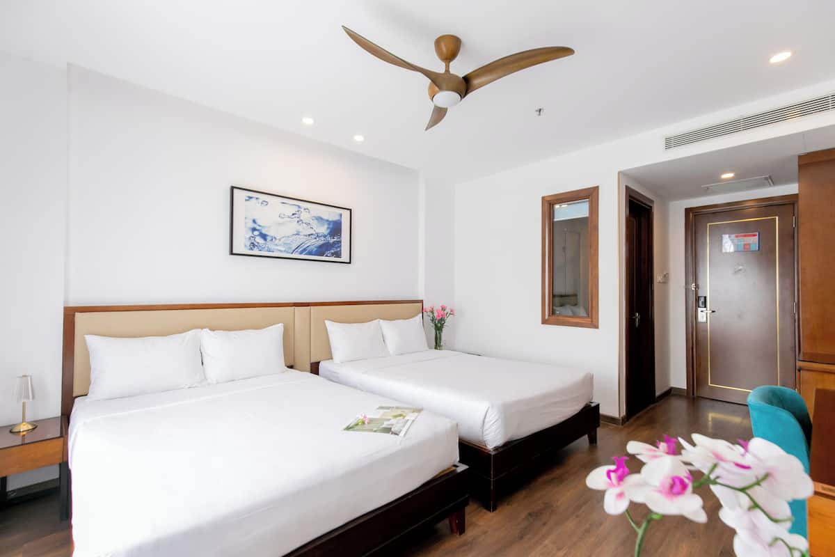 A hotel room with two beds, a ceiling fan, a framed artwork, a wooden door, and a wooden floor.
