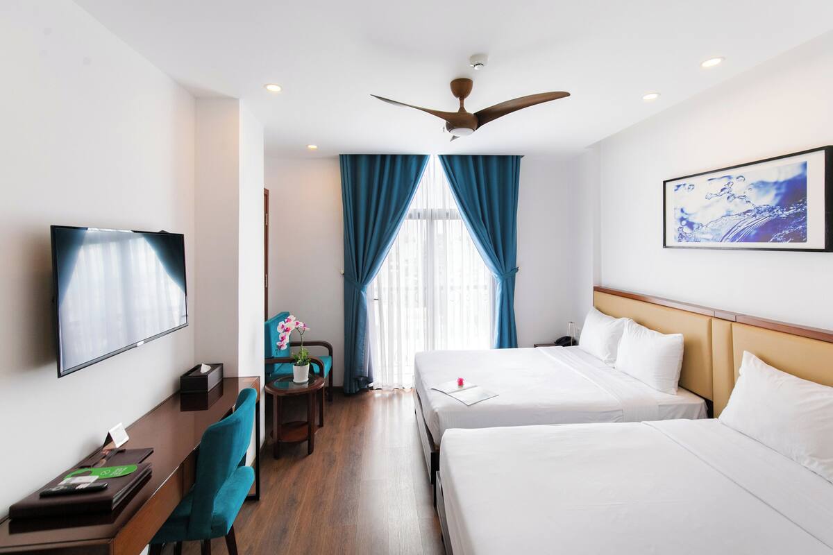 A hotel room with two beds, a desk, a chair, a TV, a ceiling fan, and a window with curtains.