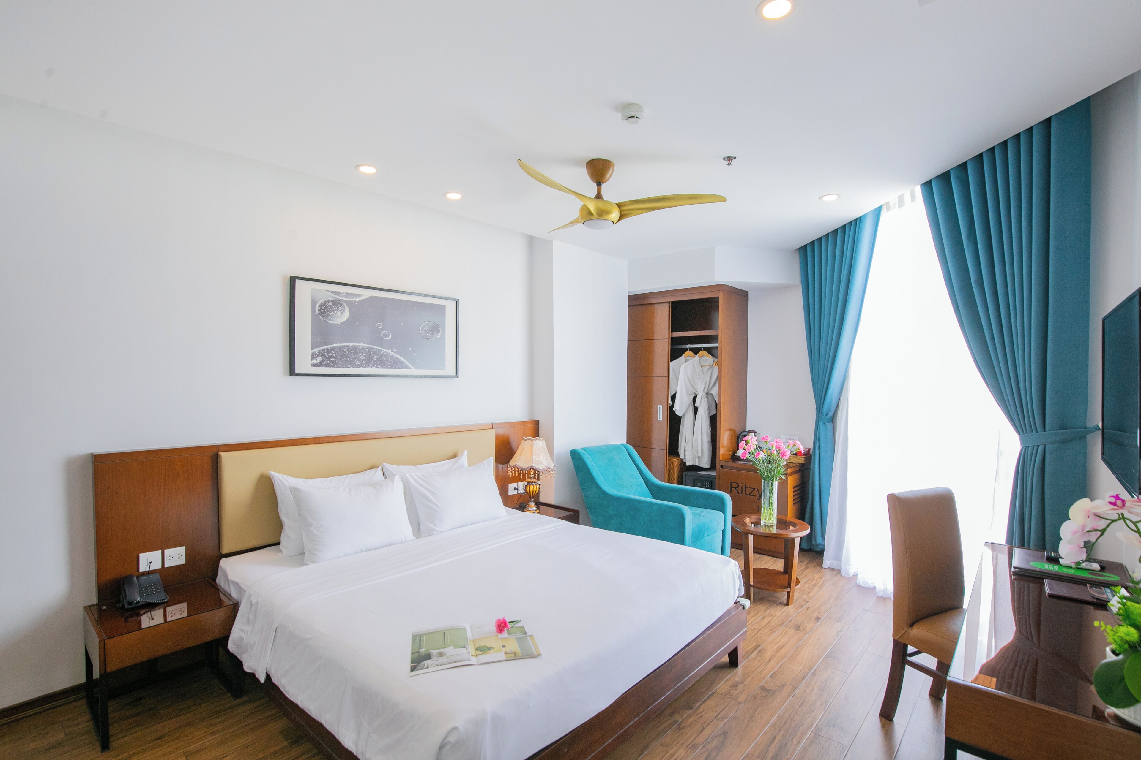 Habitación de hotel con una cama grande, un escritorio con una silla, un sillón azul, un armario y un ventilador de techo.