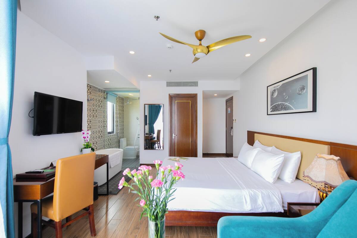 A hotel room with a bed, a desk, a chair, a TV, a ceiling fan, and a painting on the wall.