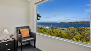 A room with a large window offering a view of the sea and boats, a black leather chair with an orange cushion, a small table with a lamp and a vase, and a dark-colored nightstand with drawers. - Manfield Seaside Bruny Island (Alonnah)