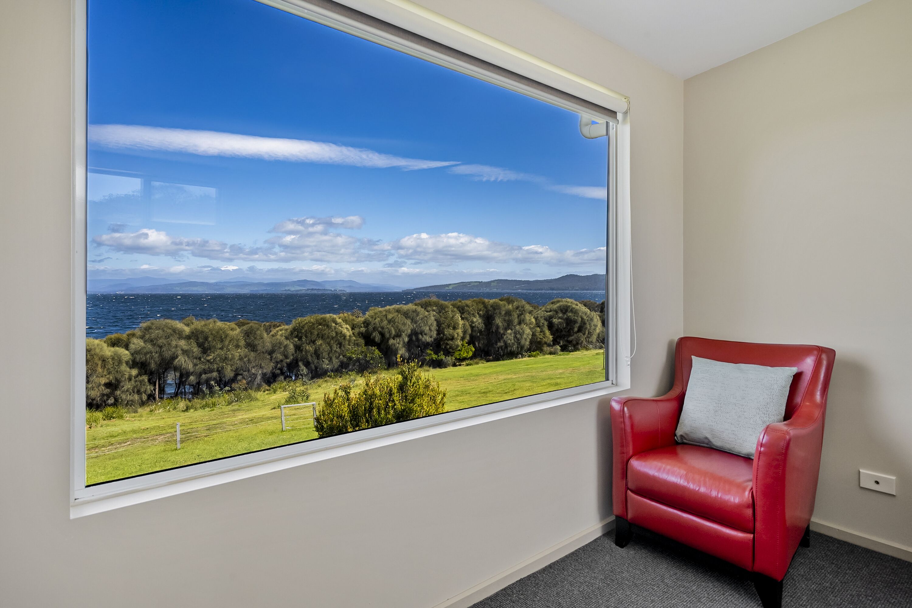 A room with a large window offering a view of a coastal landscape, a red leather chair, and a white wall.