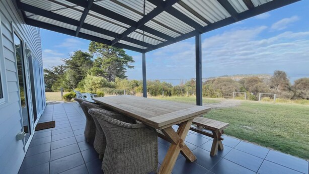 A covered patio with a wooden table and wicker chairs, overlooking a grassy area and trees. - Manfield Seaside Bruny Island (Alonnah)