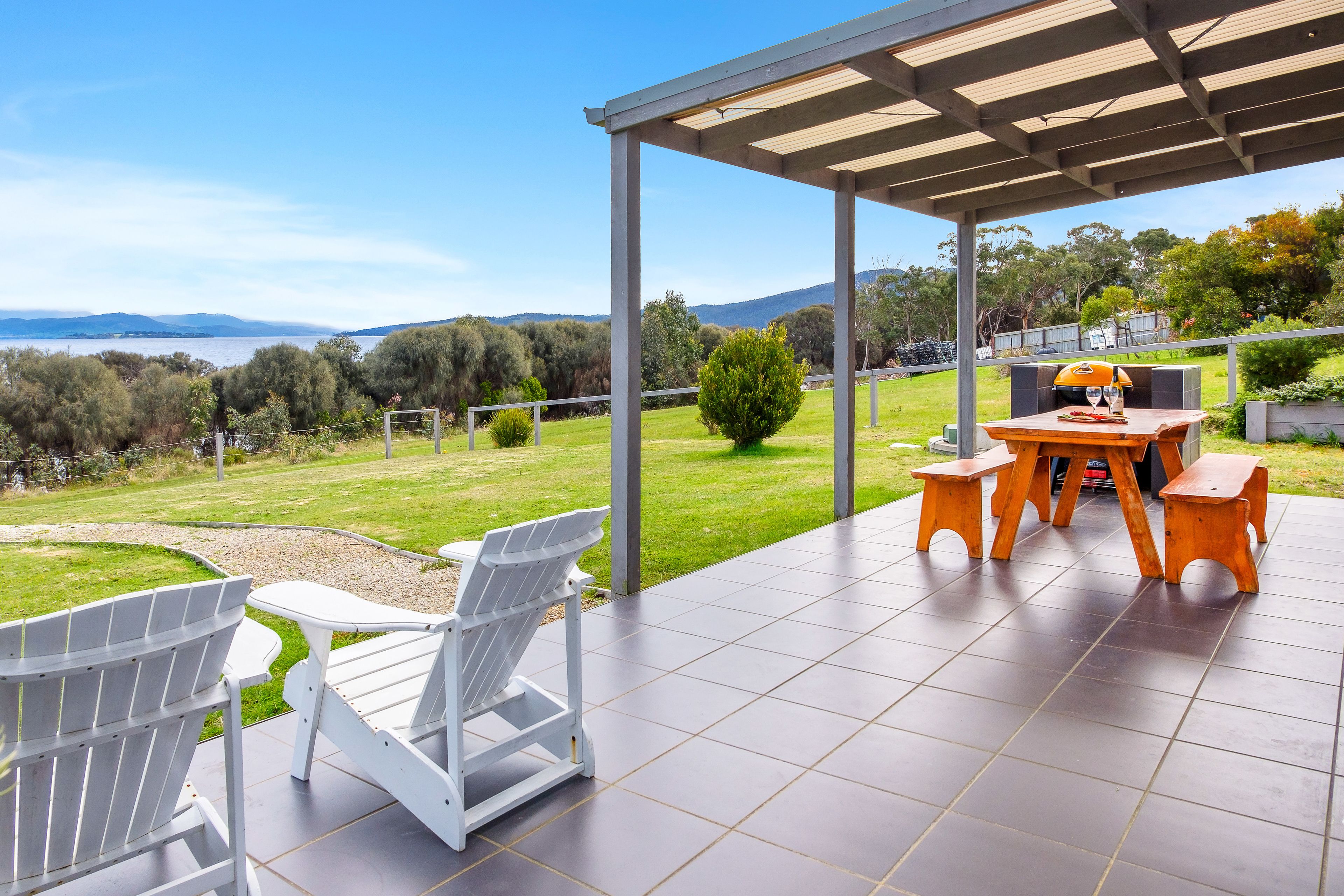 Un patio techado con vista a un lago y colinas, con una mesa y sillas.