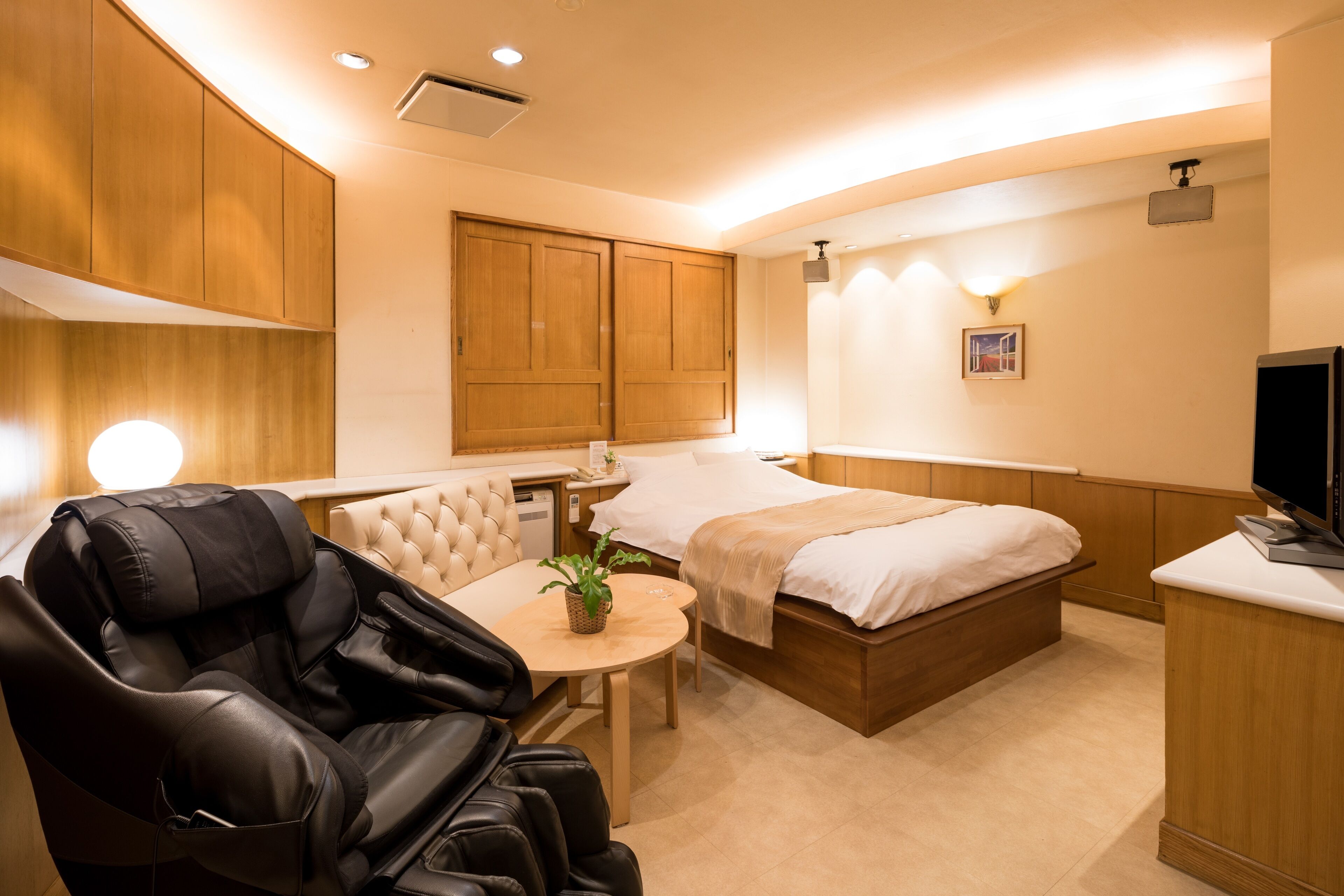 A hotel room with a massage chair, a bed, a bedside table with a plant, and a television.