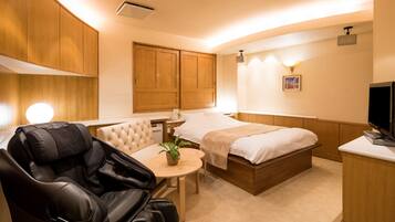A hotel room with a massage chair, a bed, a bedside table with a plant, and a television.