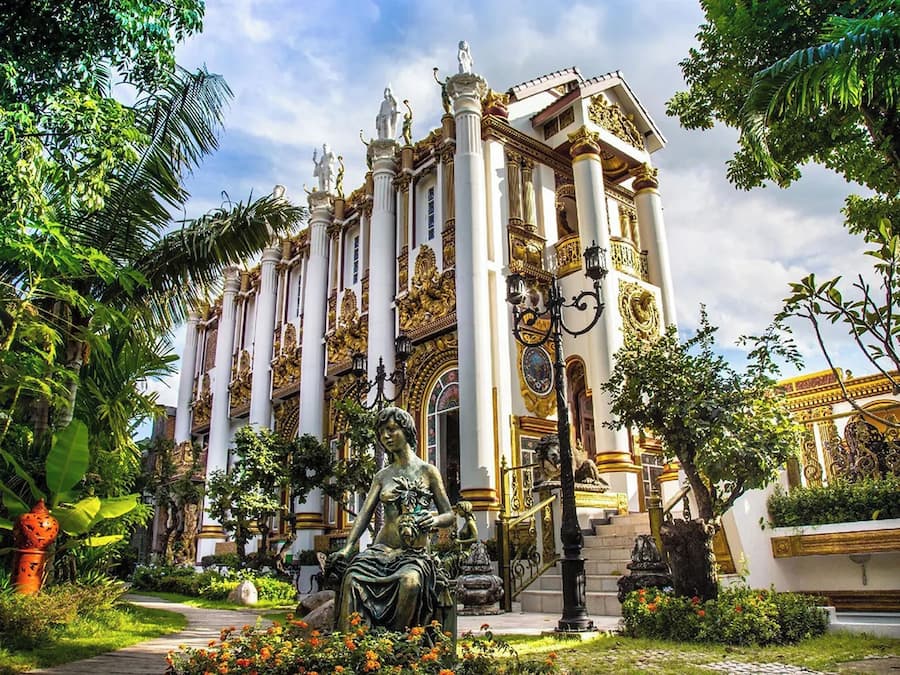 Um edifício imponente com várias colunas, uma estátua em primeiro plano e vegetação exuberante ao redor.