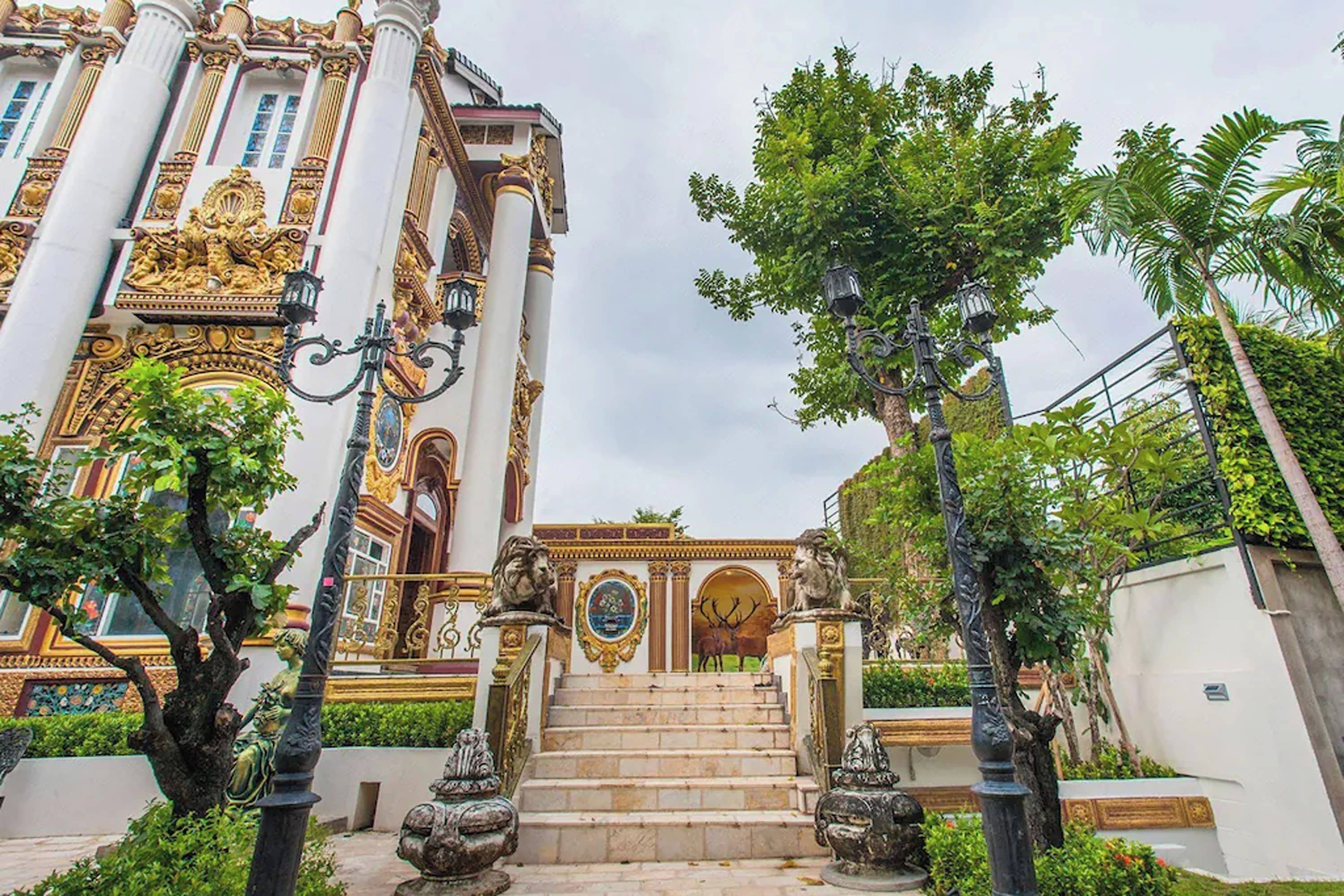 Ein prächtiges Gebäude mit verzierten Säulen und Statuen, eine Treppe führt zu einem verschlossenen Eingang, umgeben von üppigem Grün.