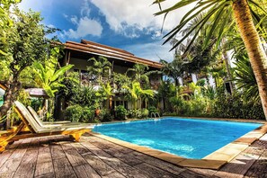 Un espace piscine doté d’une terrasse en bois, de chaises longues et d’une végétation tropicale.