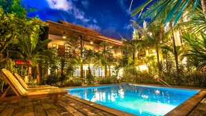 Un espace piscine doté d’une terrasse en bois et de chaises longues, entouré d’une végétation luxuriante et bordé d’un bâtiment doté de nombreux balcons.