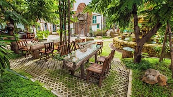 Un área de comedor al aire libre con una mesa de vidrio y sillas de madera, rodeada de abundante follaje y un suelo con diseño.