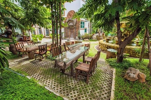 Un espace de restauration en plein air avec une table à plateau de verre et des chaises en bois, entouré d’une végétation luxuriante et doté d’un sol à motifs.