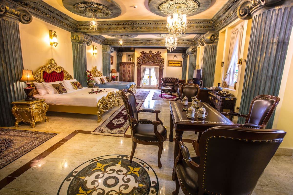 Une chambre à coucher luxueuse avec un grand lit, un coin salon avec une table et des chaises, et des décorations ornementales au plafond.