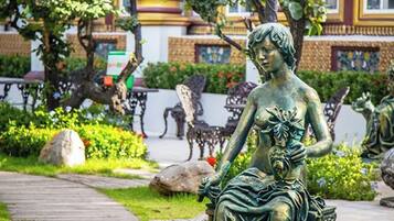 A statue of a seated woman with a flower in her hand, surrounded by greenery and flowers, with ornate architecture in the background.
