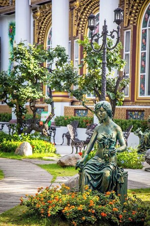 Une statue représentant une femme assise, tenant une fleur à la main, entourée de verdure et de fleurs, avec une architecture ornée en arrière-plan.