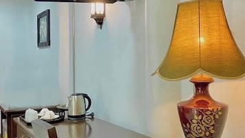 A hotel room with a flat-screen TV mounted on the wall, a table with a lamp, a kettle, and a remote control.