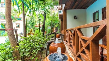 Un balcón de madera con una fuente rústica, rodeado de exuberante vegetación y un edificio con fachada azul.