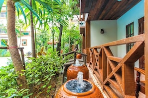 Un balcon en bois agrémenté d’une fontaine rustique, entouré d’une végétation luxuriante et d’un bâtiment à la façade bleue.