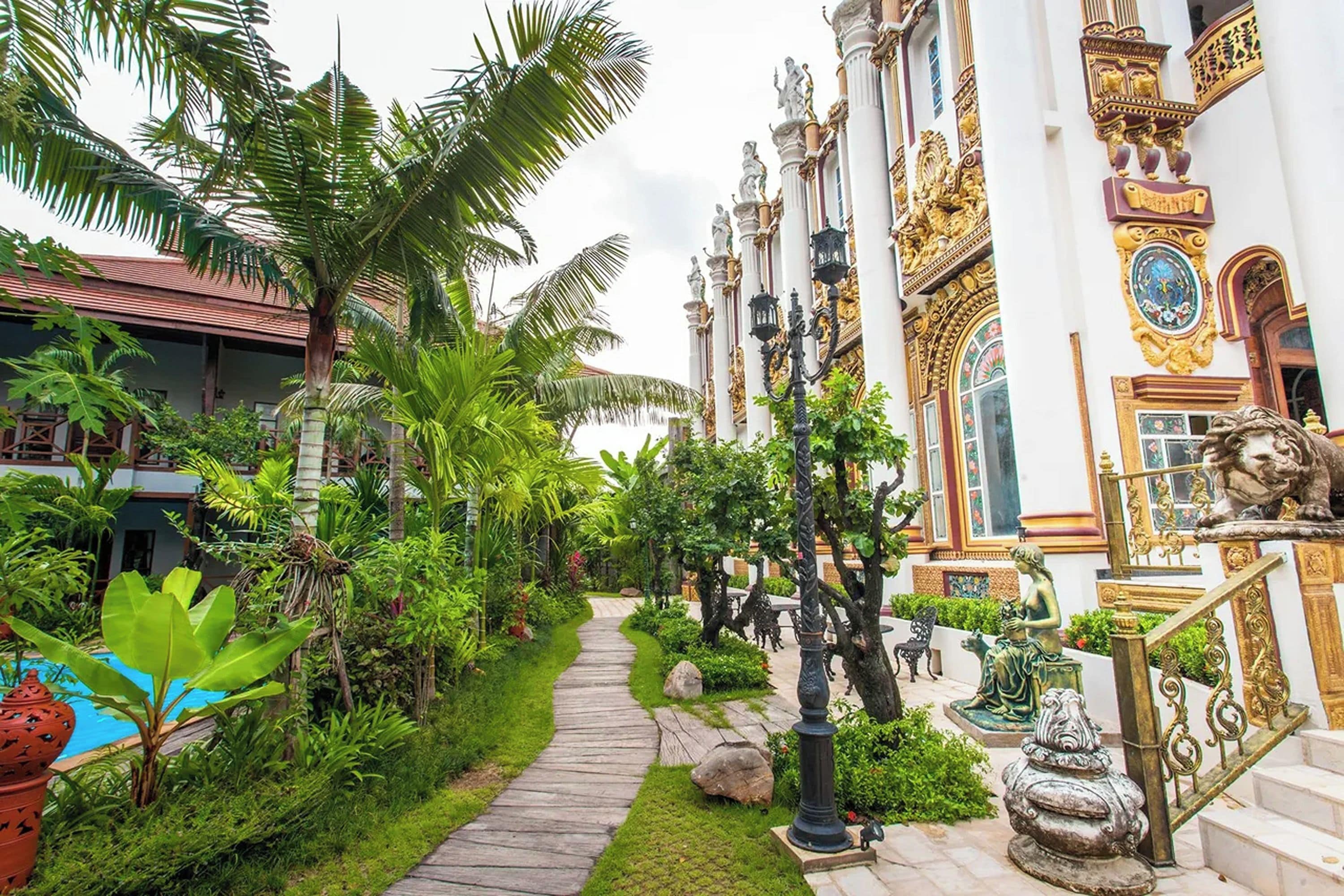 Un camino conduce a un edificio con columnas y estatuas ornamentadas, rodeado de abundante vegetación y palmeras.