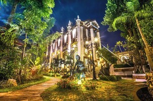 A well-lit building with classical architecture, surrounded by lush greenery and a paved pathway leading to the entrance.