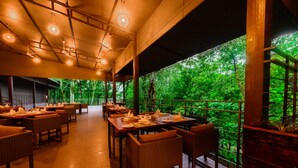 Een restaurantruimte met tafels en stoelen, grote ramen en uitzicht op groen.