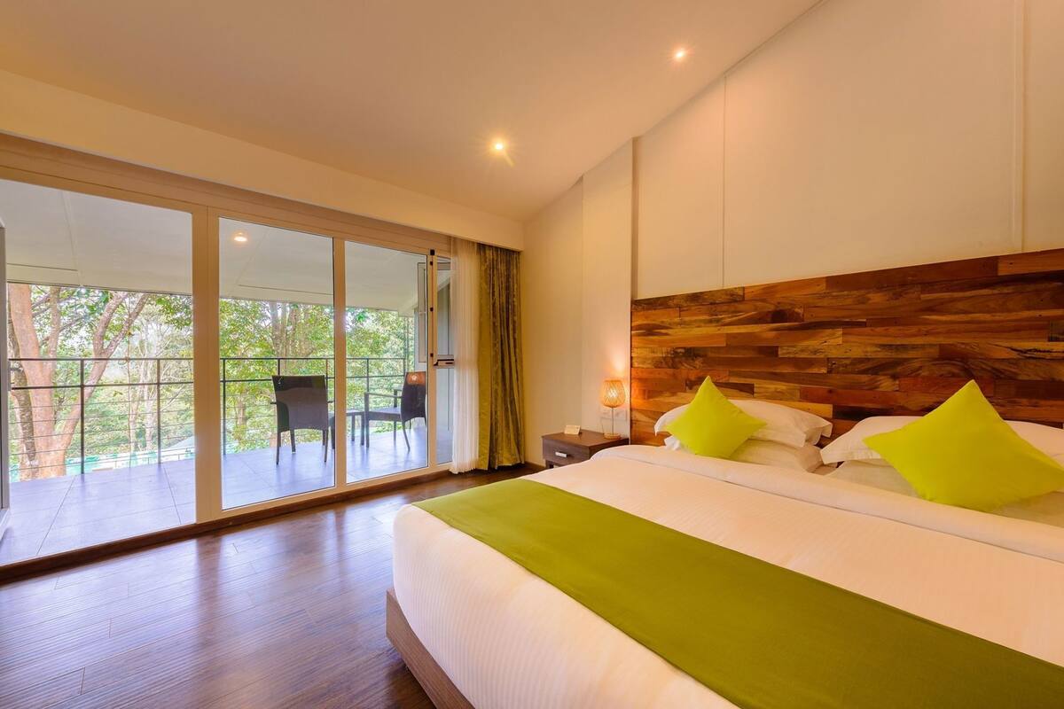 Une chambre d’hôtel moderne avec un grand lit, une tête de lit en bois, un balcon avec des chaises et une vue sur les arbres.