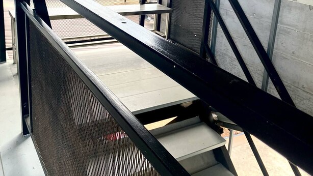 A staircase with metal railings leading to an outdoor area with a view of buildings and palm trees. - Ploynam Homestay (Amphawa)