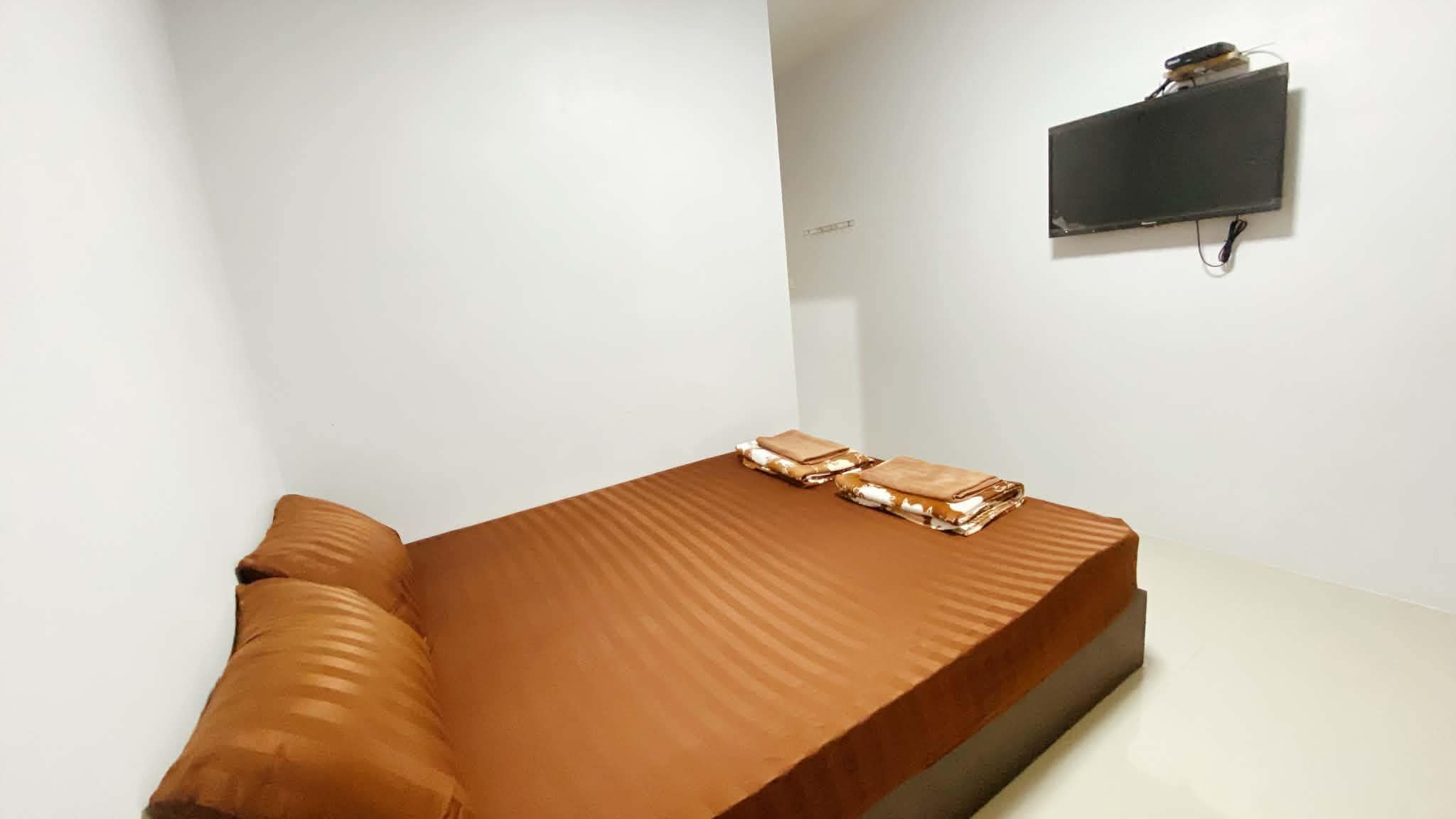 Un lit simple avec un matelas et des oreillers marron, un téléviseur à écran plat fixé au mur et une petite table sur laquelle est posée une serviette pliée.